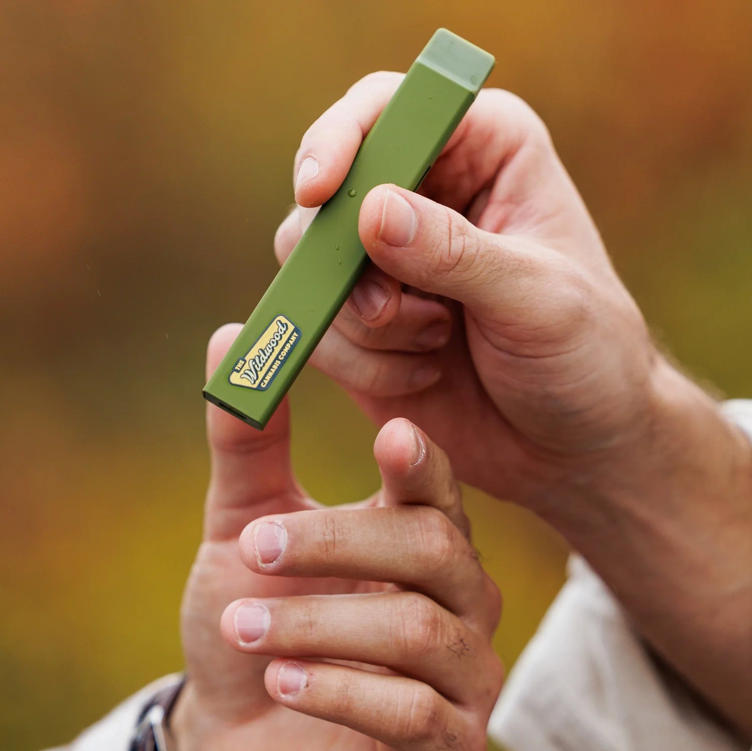 A smiling woman in a light jacket stands by a car on a rainy, tree-lined road, holding her green water bottle and Woodstock Hemp Co's Woodstock THCa Liquid Diamond Live Resin 2.0g Disposable amid autumn leaves.
