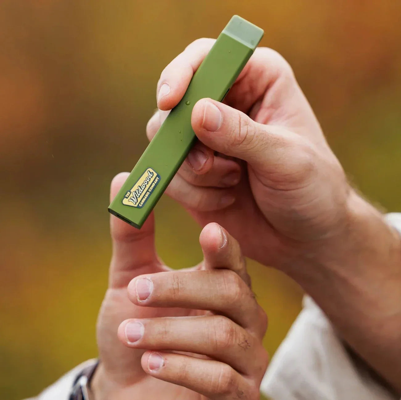 A smiling woman in a light jacket stands by a car on a rainy, tree-lined road, holding her green water bottle and Woodstock Hemp Co's Woodstock THCa Liquid Diamond Live Resin 2.0g Disposable amid autumn leaves.