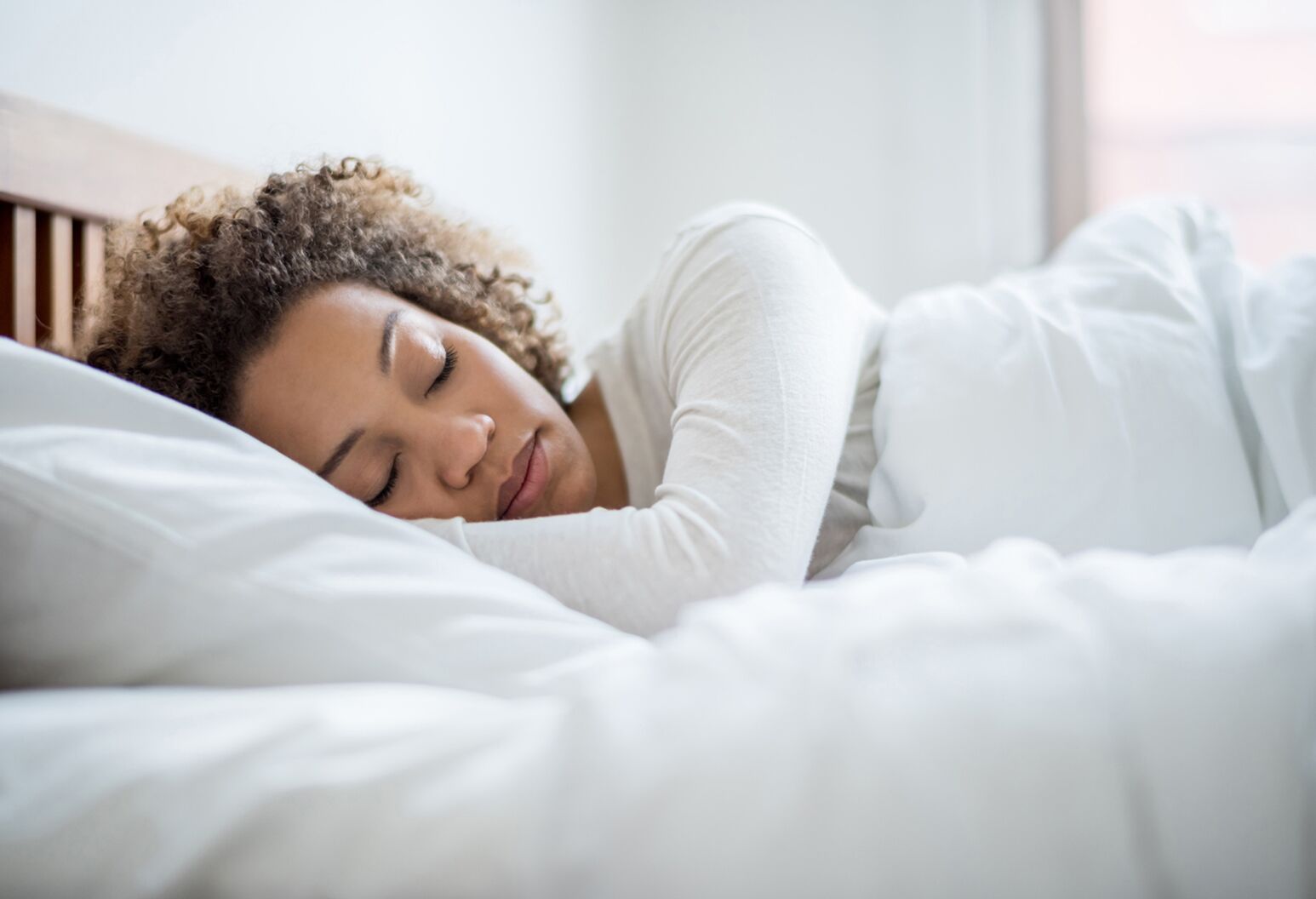 A person with curly hair peacefully sleeps on their side in a white bed, possibly enjoying the calming effects of Realize Bedtime Gummies Delta 9 + CBN by Realize, in a bright and serene bedroom.