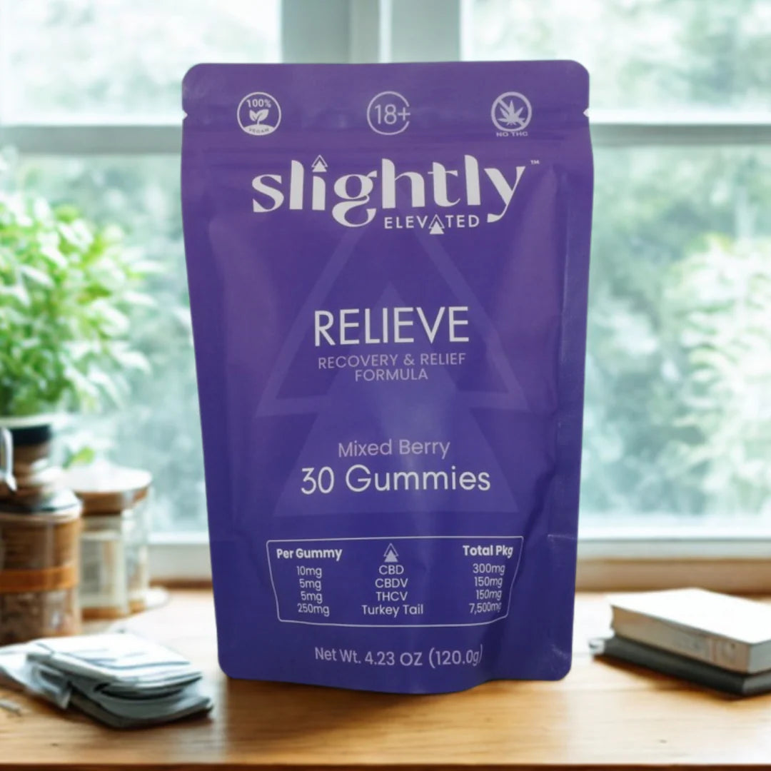 A purple pouch of Slightly Elevated Relieve Gummies 30ct stands on a table, displaying dosage, ingredients, and CBD recovery info. Books and a jar are in the background near a sunlit window.